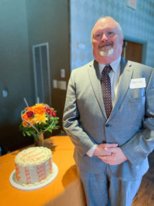 Patrick Dunn standing next to orange and white round decorated retirement cake and smiling.